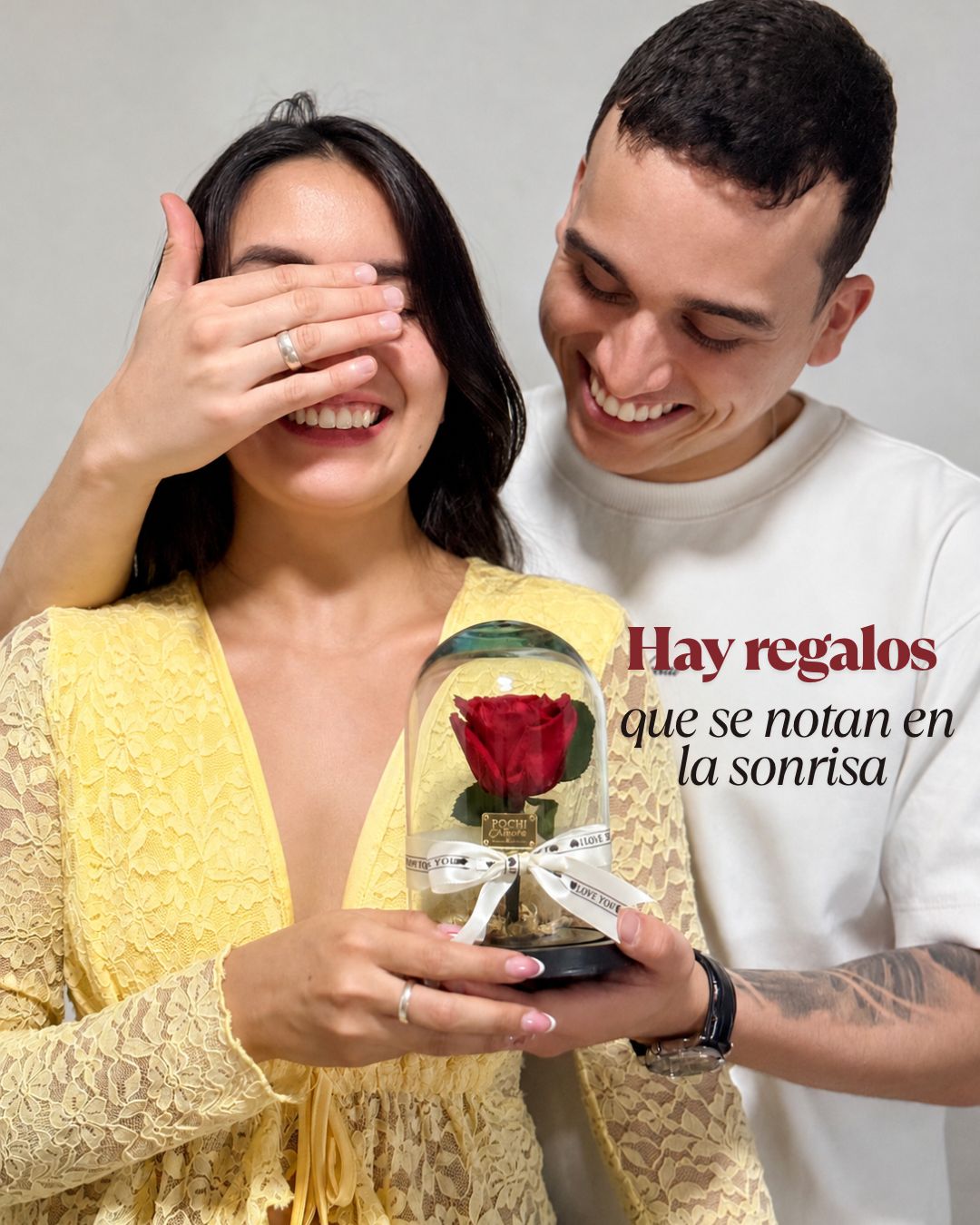 Pareja sonriendo: él le cubre los ojos mientras ella sostiene una rosa preservada en cúpula de Pochiamore